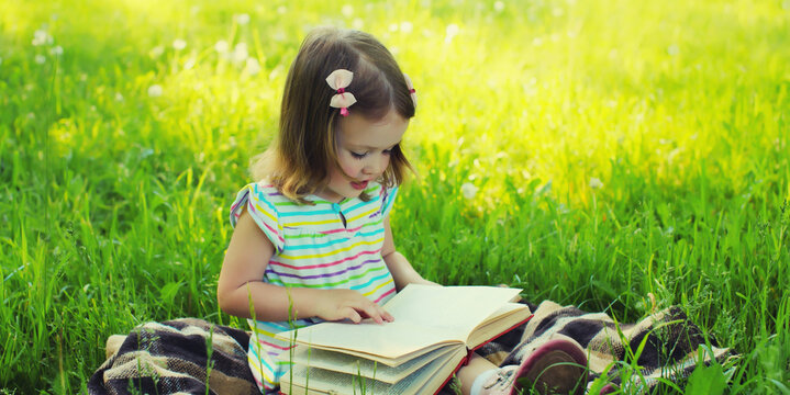 Happy little girl child reading book sitting on the grass in summer park