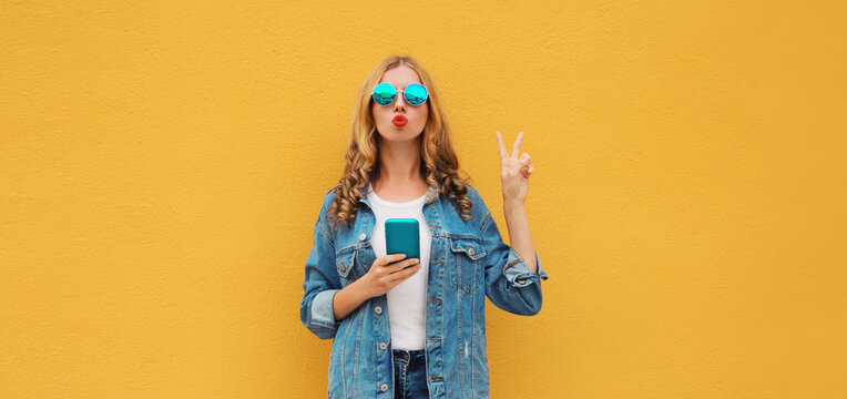 Stylish woman posing blowing a kiss with smartphone in denim jacket casual clothing posing on yellow