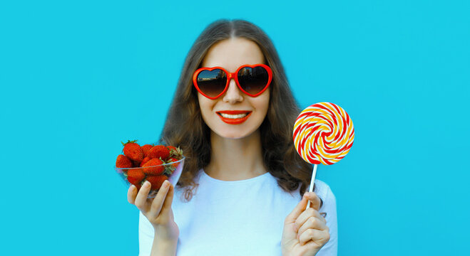 Portrait of beautiful caucasian young woman with handful of fresh strawberries holding sweet lollipop wearing red heart shaped sunglasses on blue studio background