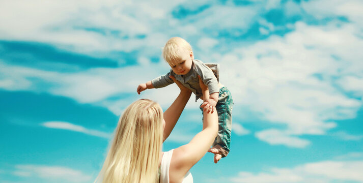 Happy young mother holding her baby raising up, joyful mom and little son child on blue sky
