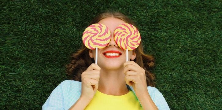 Happy young woman holding colorful lollipop, cheerful teenager girl lying on grass in summer park