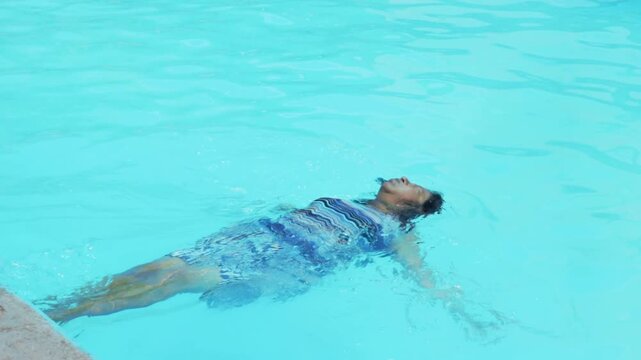 Middle-aged woman with her eyes closed falling backward into the clear blue water of a swimming pool, floating peacefully on her back while wearing a summer dress