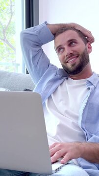 Thoughtful young man with a beard sitting on a couch while working on his laptop, changing expressions from confused to smiling and thinking as he develops his project in his living room