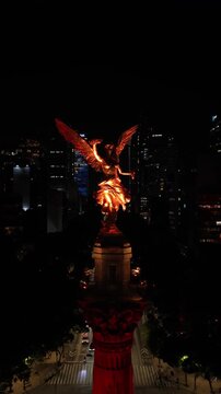 Drone hovering before illuminated Angel of Independence at night with Paseo de la Reforma skyline, vertical video, Mexico City