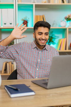 Indian man on home table smiles and waves to laptop webcam during online meeting and remote lesson. Arabian guy on home table greets screen with warm smile in video call and distance lesson at home.