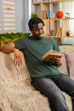 African American young man on home sofa reads book with focused look during calm leisure at home. Black guy on home sofa turns page slowly for education study relaxation in cozy living room indoors.