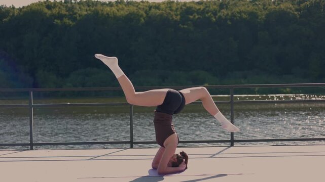 A flexible young woman performs a graceful tripod headstand pose on a yoga mat outdoors. The athletic girl balances upside down with bent legs against a scenic river and lush green forest.