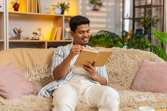 Indian man on home sofa reads a few lines in a book turns page and smiles at rest in living room. Arabian guy on home sofa enjoys quiet reading time and peaceful home leisure with warm relaxed mood.