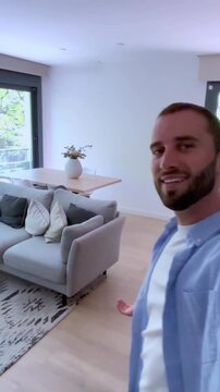 Enthusiastic young man vlogging from his modern apartment, showing the interior to his followers before sitting on the sofa, creating engaging content for his social media channel