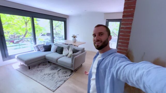 Young man holding the camera and vlogging about moving into his new modern apartment, walking through the rooms and then sitting on the sofa to talk to his followers on social media