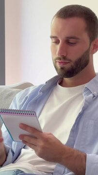 Close-up of a handsome young man with a beard sitting on a couch, thoughtfully writing down his ideas, plans, and to-do list in a small spiral notebook while studying at home
