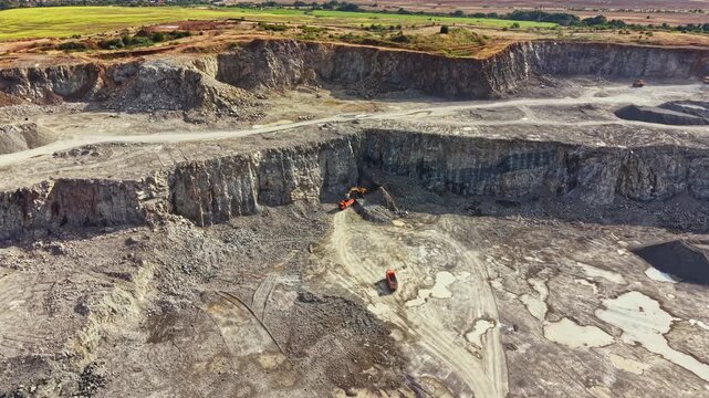 Large granite quarry with heavy machines moving dirt and rocks. The area shows uneven ground and dust from ongoing work. Sunlight shines over the quarry, revealing open space.