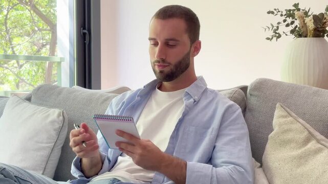 Focused handsome bearded man sitting on a comfortable couch and writing down his thoughts, plans, or a to-do list in a notepad while relaxing in his modern living room on a sunny day