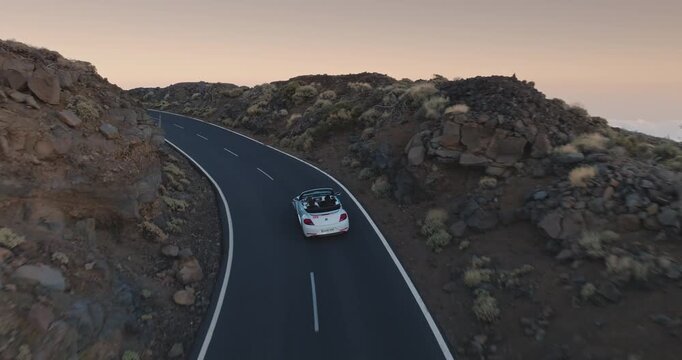 White convertible car traveling along a winding asphalt road, exploring volcanic landscape of Teide National Park, Tenerife, under bright sunset sky. Travel destination. Aerial tracking drone flight