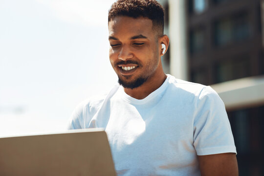 Smiling Black Male Professional Using Laptop While Sitting Outdoors With Wireless Earbuds, Engaged In Work. Modern Setting Represents Freelancing And Business Lifestyle In Urban Environment.