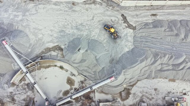 A yellow machine works in a granite quarry. The area is filled with large piles of stone gravel. Equipment is placed around the site. This occurs in a busy mining location.