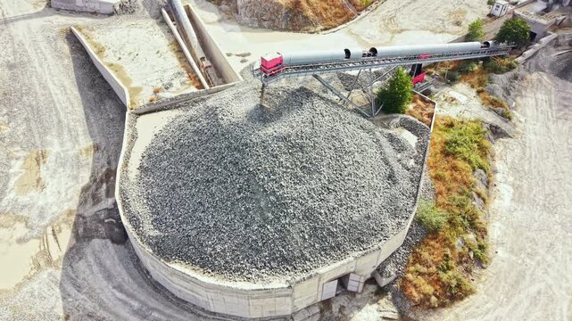 A large granite quarry shows a big pile of crushed stone. A conveyor belt moves materials from one area to another. The site appears busy with mining activity.