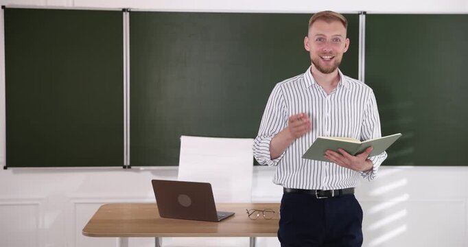 Smiling teacher with book conducting lesson near table in classroom