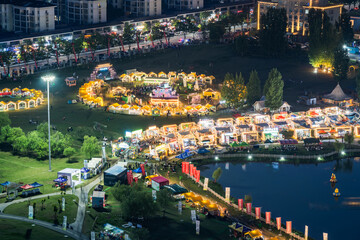 Fototapeta premium An illuminated outdoor night market and festival event set by a lake in an urban park, with stalls and event structures at night.