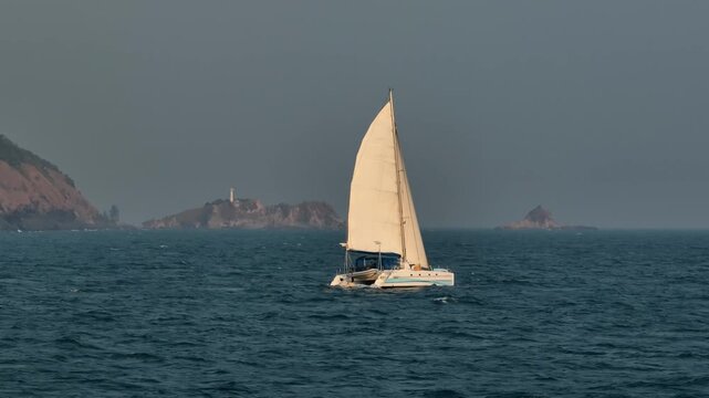 Catamaran sailboat navigating the blue sea, passing a distant island with a lighthouse under an overcast sky near the coast of Koh Lanta, serene natural scene of summer travel and Thailand exploration