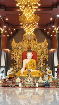 A magnificent golden Buddha statue seated inside an ornate Thai Buddhist temple, surrounded by intricate golden decorations and elegant chandeliers. A peaceful spiritual interior reflecting Thai cultu