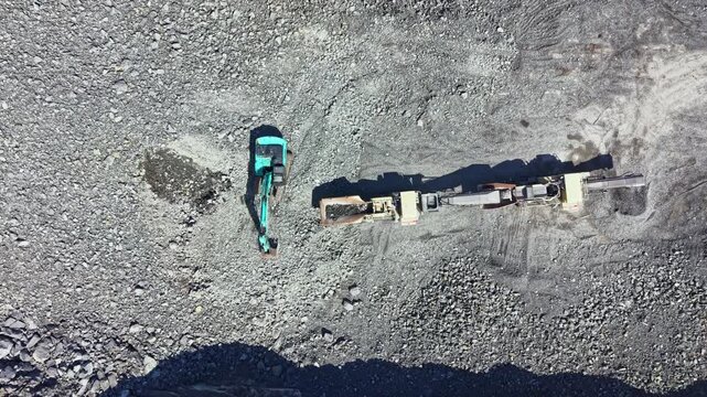A machine works at a granite quarry. An excavator digs into the ground while a large truck waits to transport the stone. The site is busy with rock and dust surrounding the area.