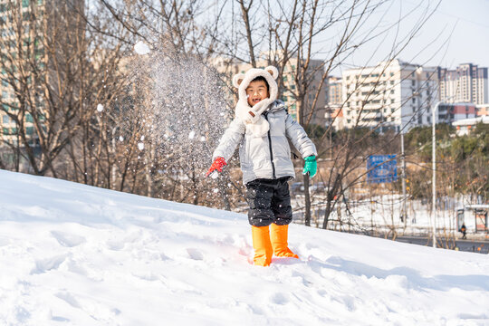 A young child in warm winter clothing plays with snow on a snowy urban hill outdoors on a clear cold day.