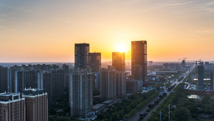 Obraz premium Aerial view of modern urban skyscrapers under a bright sunrise sky with a city road and green spaces.