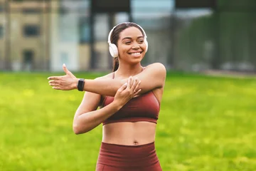 Smiling Asian Woman Stretching Arm While Listening To Music With Headphones In Park, Engaged In Outdoor Fitness Routine, Enjoying Active Lifestyle Surrounded By Nature. Weight Loss Concept. © Prostock-studio