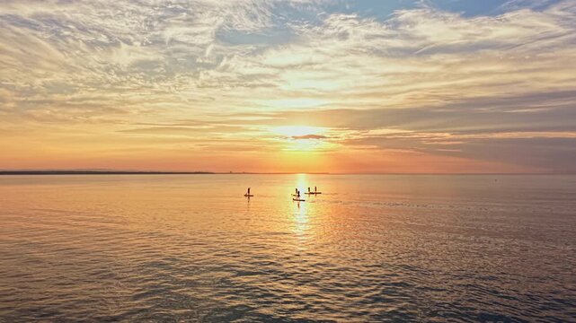 Groups of paddle boarders glide across the water as the sun sets in the background. Colors of the sky change to orange and yellow. The scene shows a peaceful evening by the shore.