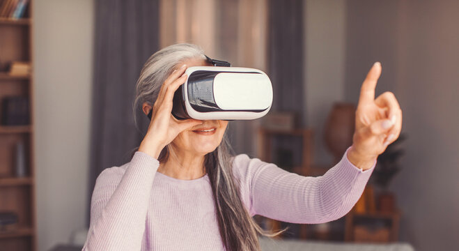 Focused Senior Caucasian Woman Interacting With Virtual Reality Headset At Home, Enjoying Innovative Technology Experience, Engaging In Immersive Digital Interaction In Comfort Of A Modern Living Room
