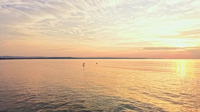 The sun sets over a calm water body, casting orange and pink hues across the sky. Two surfers stand on their boards far away, enjoying the evening.