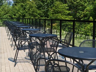 Fototapeta premium Rows of dark metal tables and chairs sit empty on a paved terrace. Green trees frame a black railing beyond the patio. The sun glints off the tables' smooth surfaces. No people occupy the space