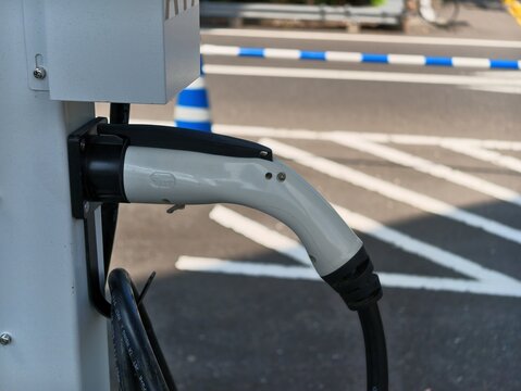 Electric vehicle charger plugged into a station. White and black cable rests against grey wall. Blue and white barriers mark parking zone. Sunlight casts clear shadows on asphalt