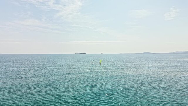 Two people are enjoying windsurfing on the water under a clear sky. Distant ships are visible on the horizon. This scene takes place during summer.