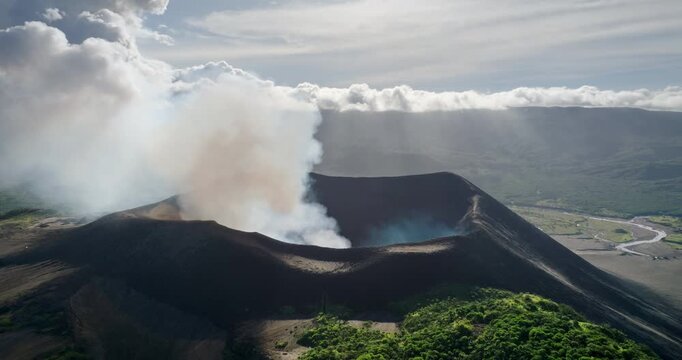 Mountain Yasur, an active stratovolcano erupting with large plumes of smoke rising from its crater, cloudy sky with green hills and a winding river. Explore Tanna Island, Vanuatu volcano. Aerial shot