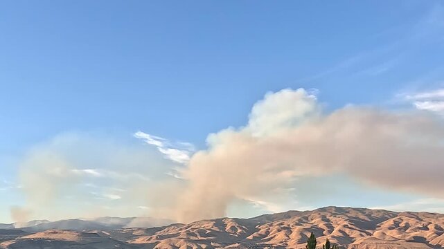 Smoke plume from a wildfire slowly rising over dry mountains under a clear sky.