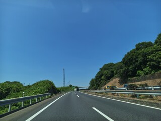 Fototapeta premium Clear blue sky stretches above winding asphalt road. Green trees line both sides, offering natural shade. Railing guards the edge, ensuring driver safety