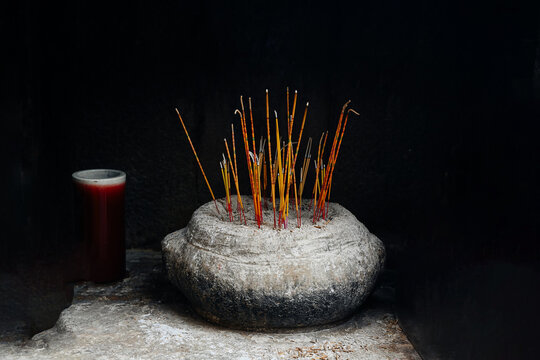 Burning traditional joss incense sticks in a rustic stone burner against dark black background inside chinese temple