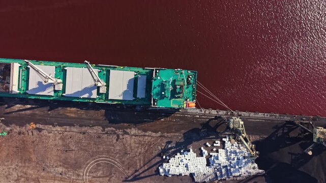 A cargo ship is positioned at a river dock, unloading large materials. Workers are seen on the ship and along the shore. The scene captures a busy time of day with ongoing operations.