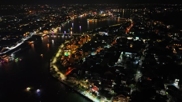 Aerial perspective of brightly lit waterfront houses and boats on the river at night in Hoi An, Vietnam, highlighting the lively atmosphere and reflections