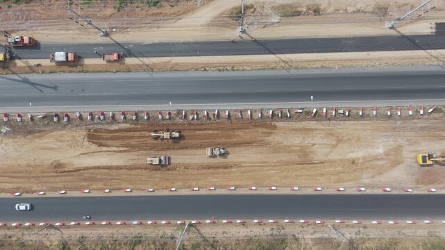 Aerial top down footage of an active road construction site with heavy machinery, passing vehicles, and earthworks. For infrastructure, transportation, and urban development concept