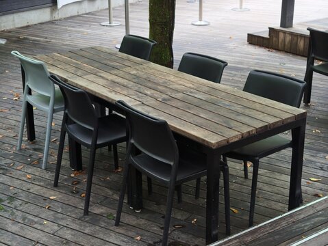 Weathered wooden table with six modern chairs. Black and gray chairs surround rustic table. Wooden deck features fallen leaves and tree trunk. Natural light enhances casual outdoor dining setup