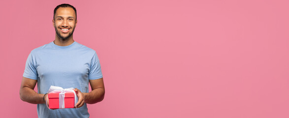 Obraz premium Smiling Arab Man In His 20s Holding A Gift Box With A Ribbon In Front Of Pink Background, Expressing Joyful Emotion. Engaged In Celebratory Activity In A Festive Setting. Celebration Concept.
