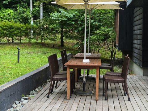 Wooden table with chairs under a yellow umbrella. Deck overlooks green lawn and trees. Quiet outdoor dining spot feels calm and inviting. Ideal for cafe, restaurant, or garden retreat imagery