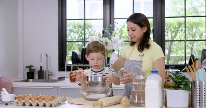 Caucasian mother guides young son sifting flour through metal sieve into mixing bowl, building cooking confidence through home baking lesson, patient parenting and family bonding in modern kitchen.