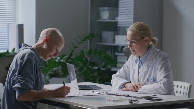 Bald female cancer patient signing medical consent form at desk and shaking hands with doctor during clinic visit