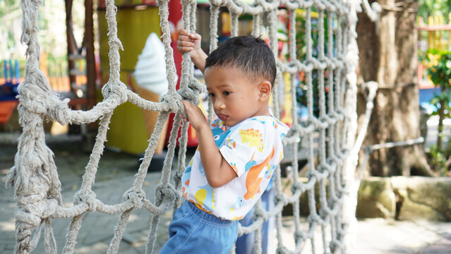 An Asian preschool boy climbs a rope net in an outdoor playground. An active child playing and trying new things, exercising in the park on a sunny day                  