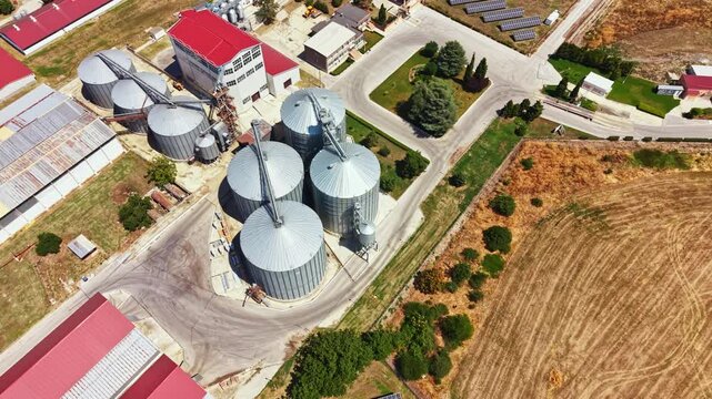 Aerial view of a pig farm showing large silos and multiple buildings used for grain storage. The farm is located in an open area with fields around.