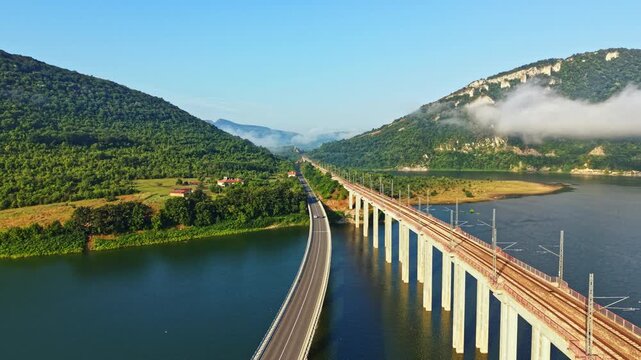 High angle view of long concrete bridge carrying vehicles across broad river toward misty valley framed by forested hills, warm morning light, soft haze, calm landscape and open horizon